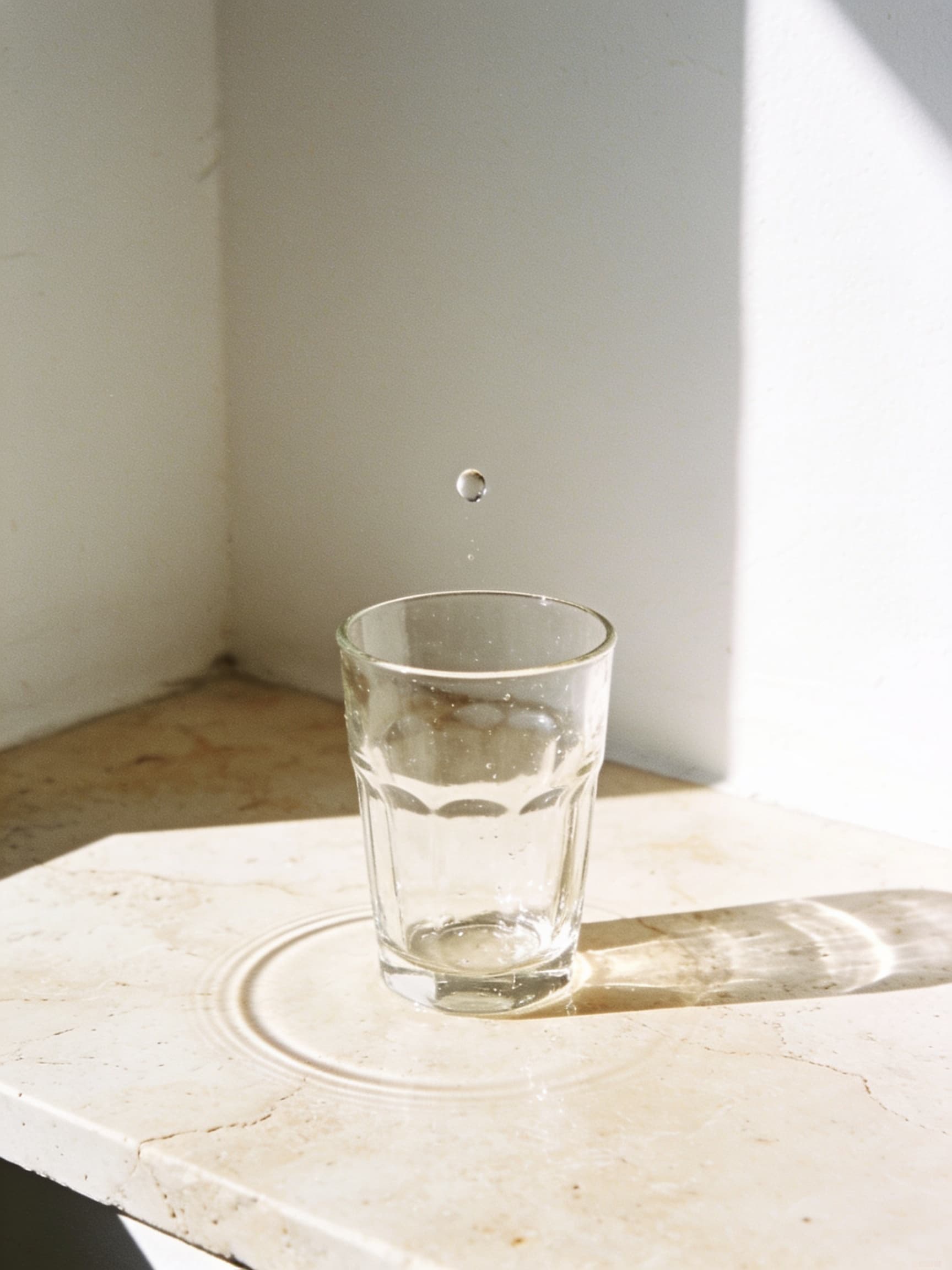A drop of water falling toward a clear glass on pale travertine in bright morning sun, a faint water ring beside it