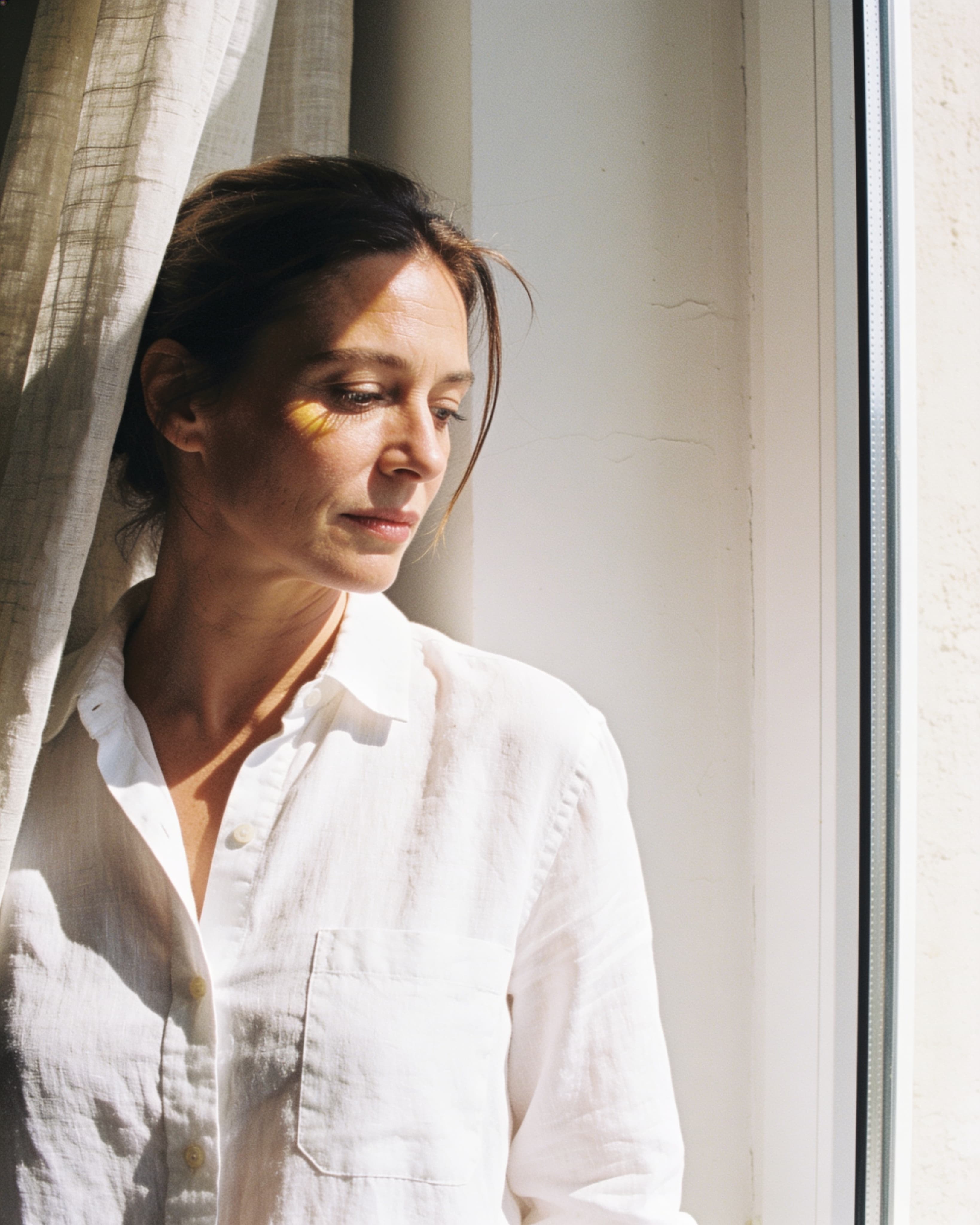 A woman at a sheer-curtained window in bright morning light, her face turned into the sun, the studio soft behind her
