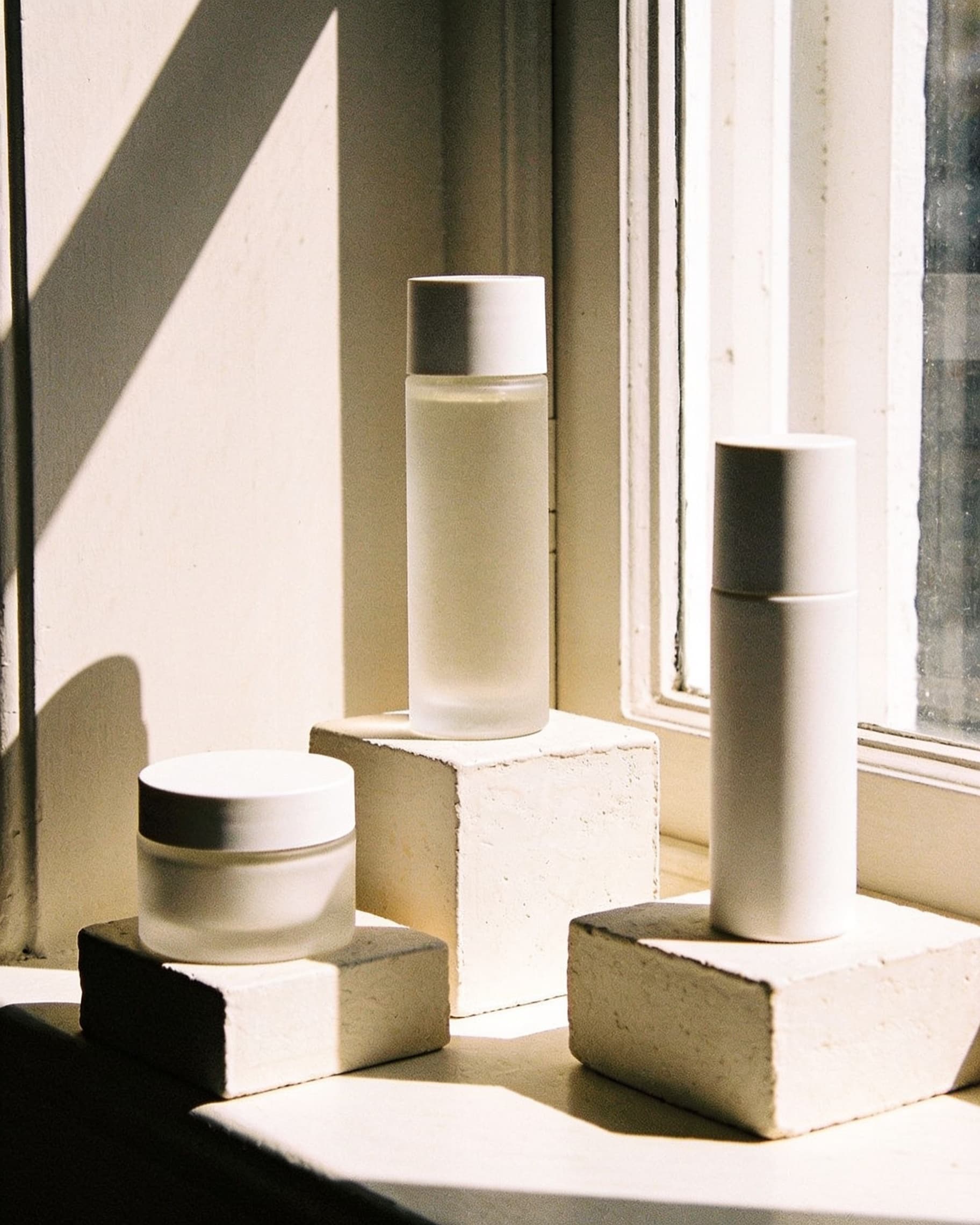 Unlabeled skincare vessels on a sunlit sill, clear and amber glass in the morning window