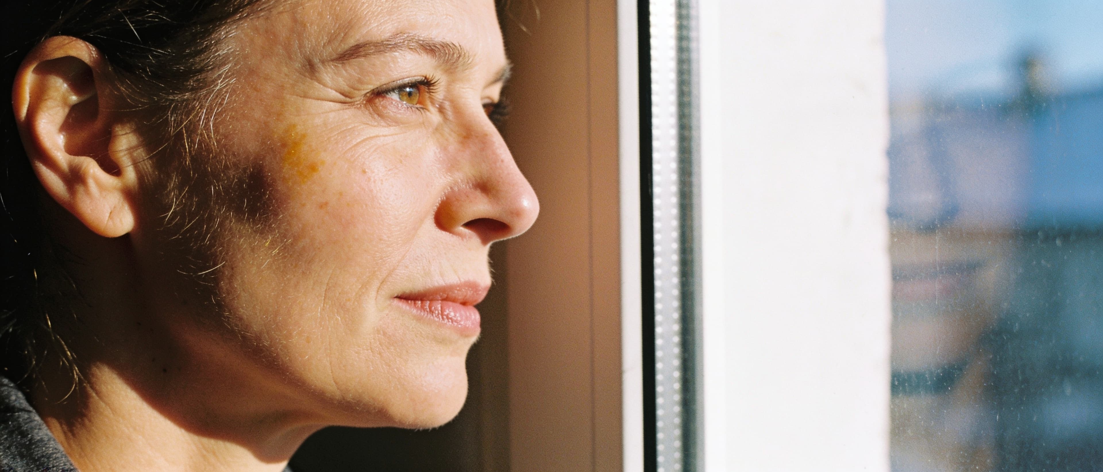 Daylight profile of a woman in her late forties by a bright window — fine lines and real skin texture in soft morning light
